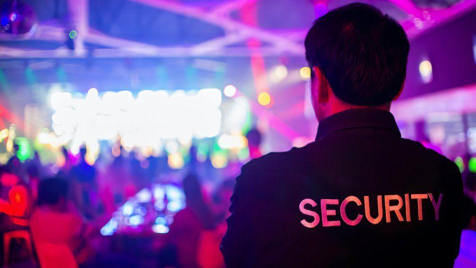 The back of a male security guard watching people inside a club. There are pink and blue lights and a stage in front of him. He is wearing a black t-shirt which says Security across the back.