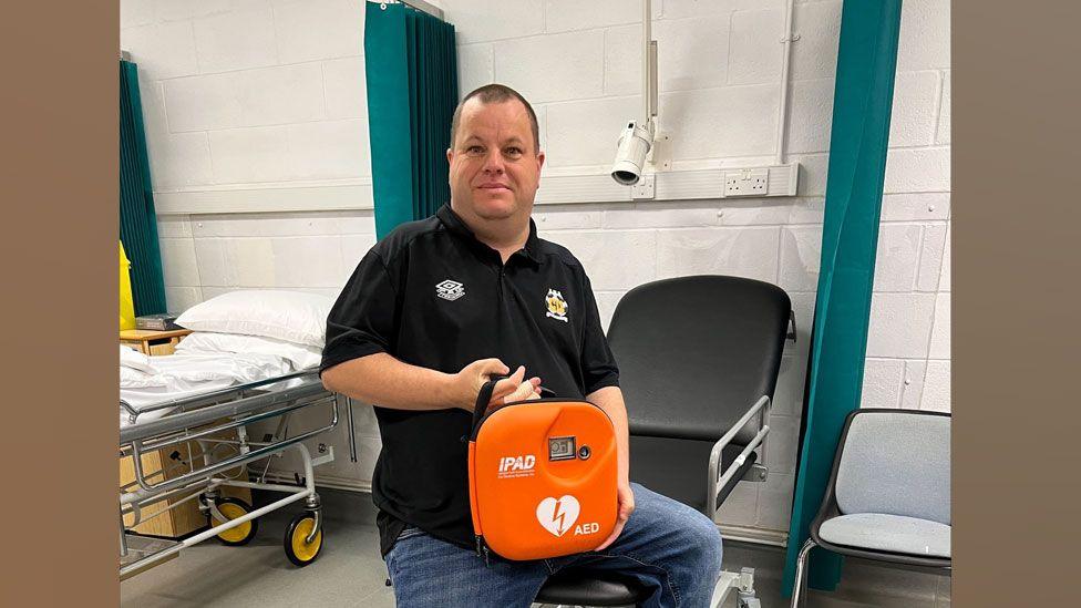 Paul Harrison wearing a black polo-shirt and perched on a black hospital trolley, holding an orange box containing an automated external defibrillator. He has short dark hair and is looking at the camera. 