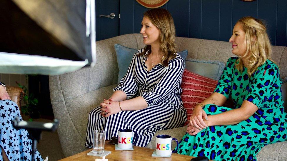 Two women sitting on a sofa. Ms Middleton-Hughes is sitting on the left and wearing a stripy dress and Ms Murray is wearing a spotty dress