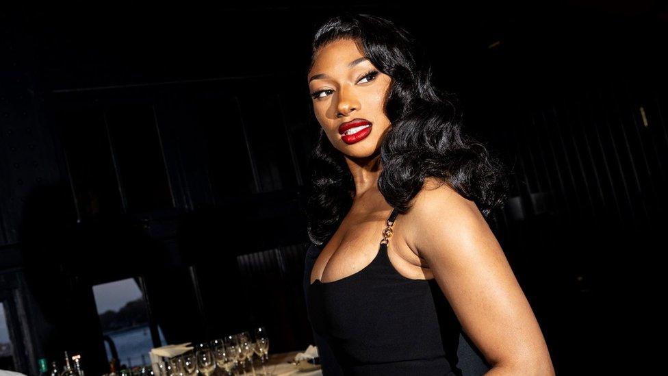 Megan Thee Stallion at a gala event in New York on 16 April 2024. Megan is a 29-year-old black woman with long wavy black hair. She wears a low-cut black dress and a bold red lipstick. She's pictured from the side, looking just past the camera over her left shoulder. The room behind her the room is dimly lit and there are champagne glasses laid out on a table.