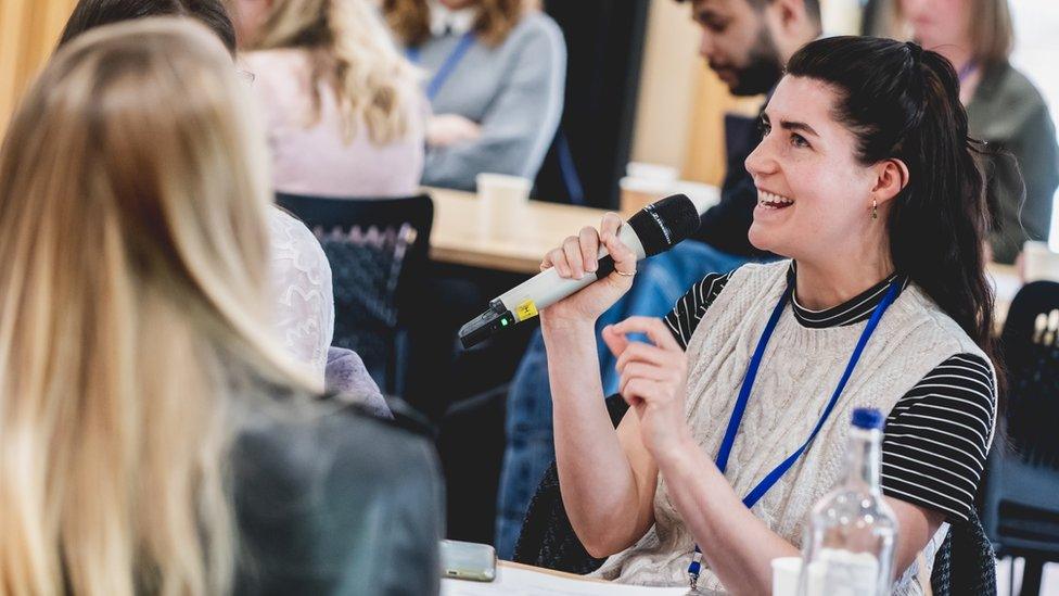 Woman with long black hair is holding a microphone and talking to students