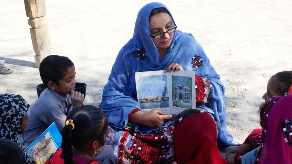 Children listen as a woman reads a book brought by a camel in Mand, Pakistan