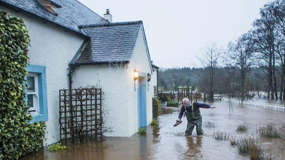 A man holds a pair of shoes as he wades through flood water in Straiton, Scotland,