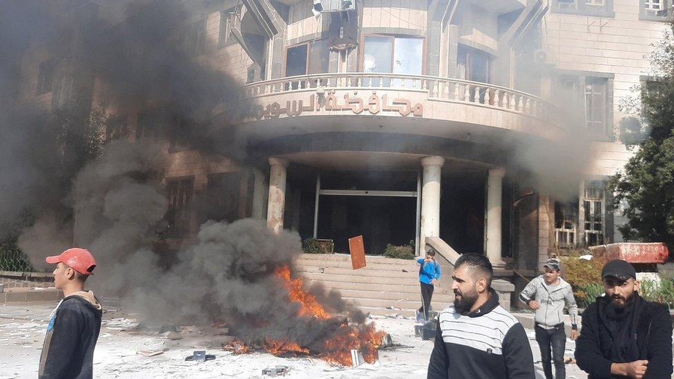 Smoke rises near a building as people take part in a protest in Suweida, Syria, (4 December 2022)