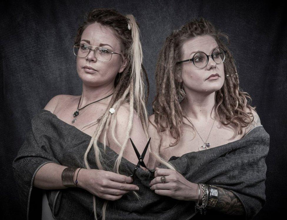 Two women wearing matching grey shirts stand back to back while holding a pair of scissors