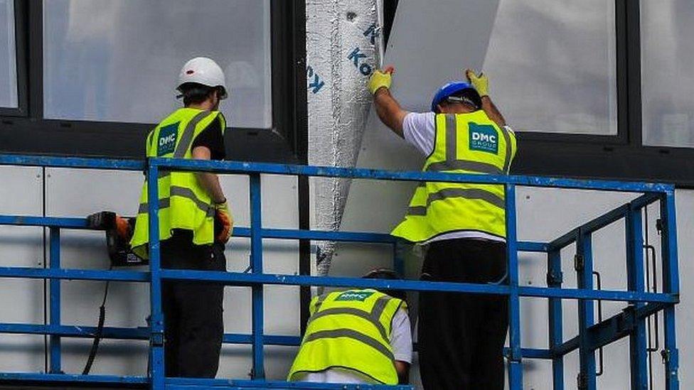 Three men in yellow hi-viz remove a panel from a block of flats