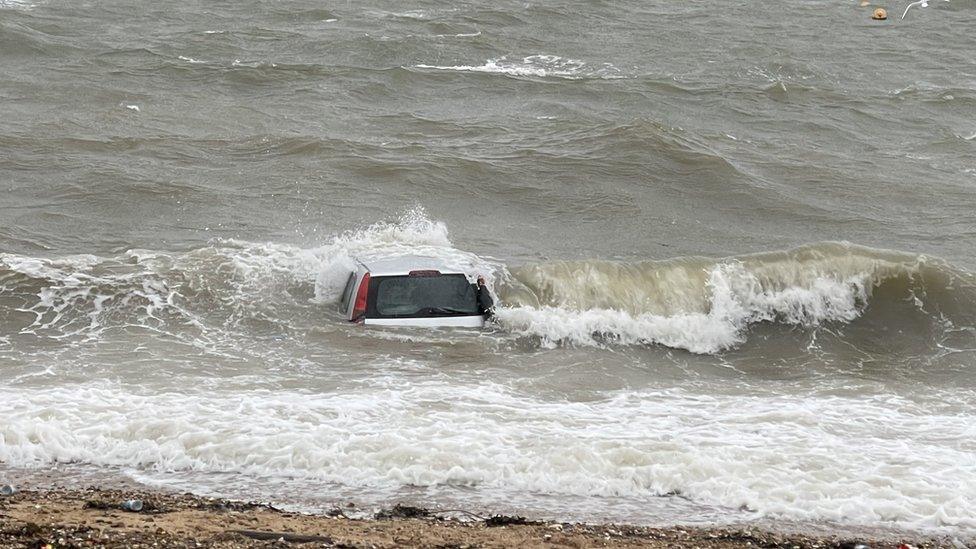 The silver hatchback's roof and rear window are visible among the crashing waves, a few feet out to sea