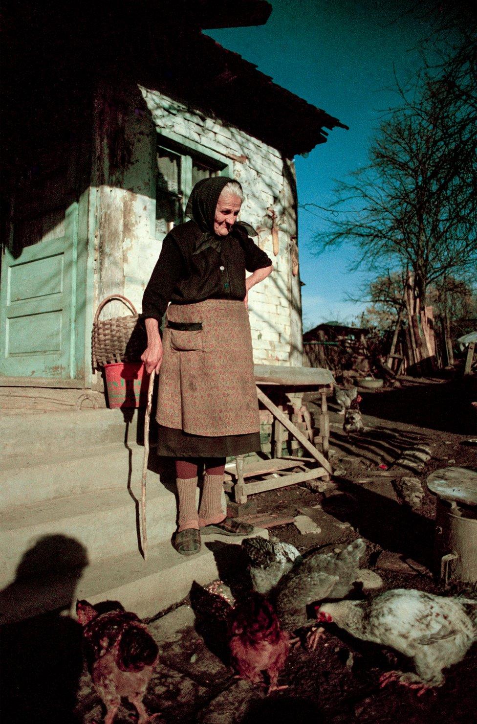 An elderly woman looks at her chickens outside of her home