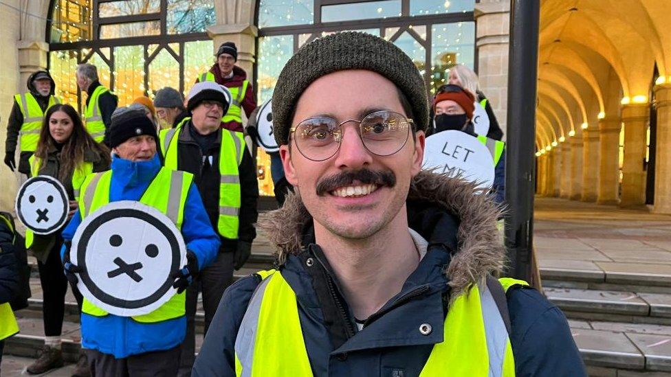 Man with woolly hat, hi-viz and moustache stands on Guildhall steps