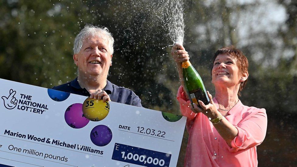 Lottery winners Michael Williams, holding a giant cheque, and Marion Wood, spraying champagne from a bottle, celebrate their £1m lottery win