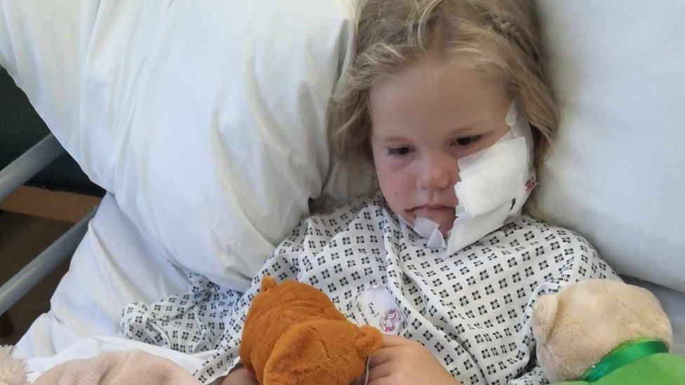 A girl lies in a hospital bed with a large bandage across her cheek