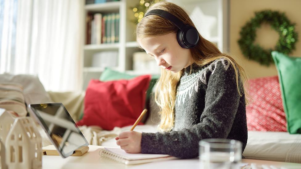 A young woman writes while listening on headphones with a Christmas wreath in the background of the living room