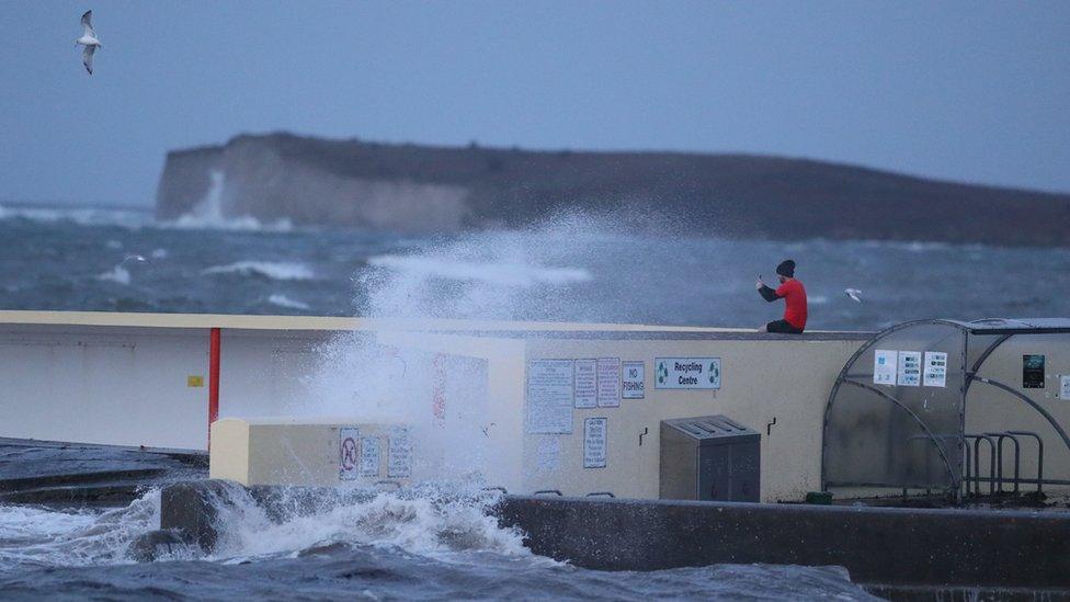 A man sits on a wall while waves crash around him