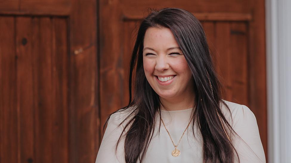 A woman with long, straight black hair smiles widely, her eyes scrunched up as she appears to laugh. She's wearing a white top, gold pendant and stands in front of a large wooden door polished to a shine.