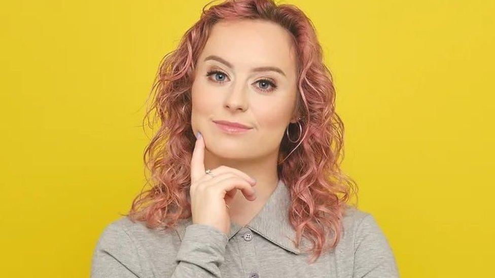 Dani Johns with her finger on her chin stood in front of a yellow background