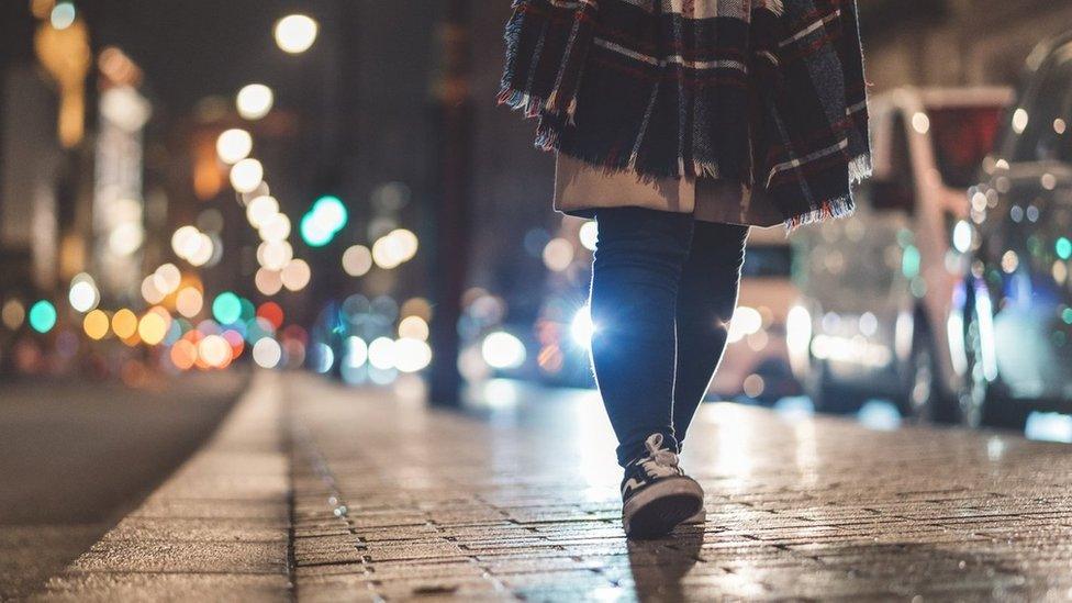 A woman walking at night along a street lit by street lamps