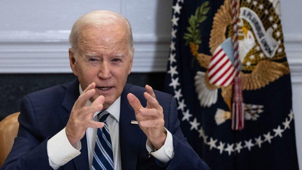 Joe Biden sitting with hands raised, before flag