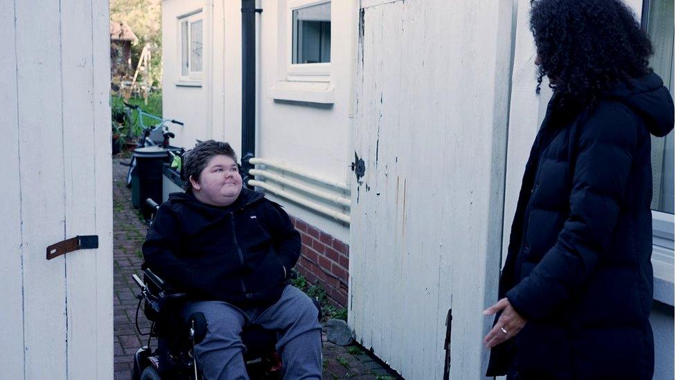 Seth in his electric wheelchair at the side entrance to his house with Nina