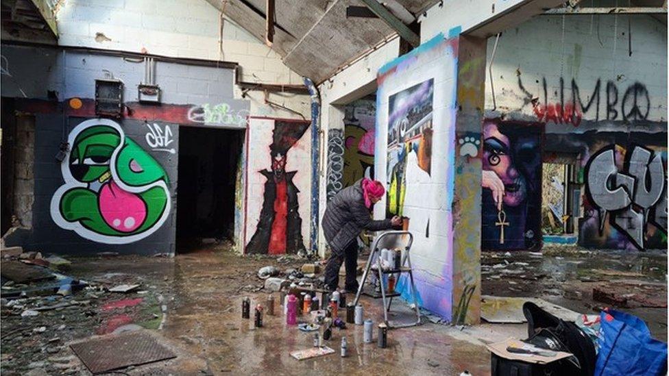 Ms Harris in a derelict building surrounded by spray cans painting the front cover for "Contraband: Bad Benny" on a wall in Stroud