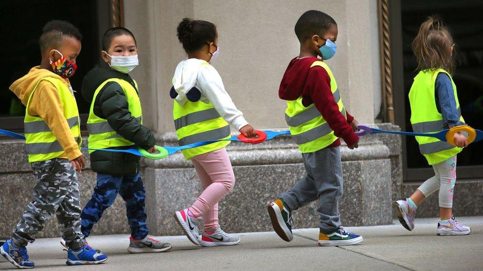 Youngsters in a preschool class wear vests and masks and hold on to a strap as they take a walk outdoors in Boston