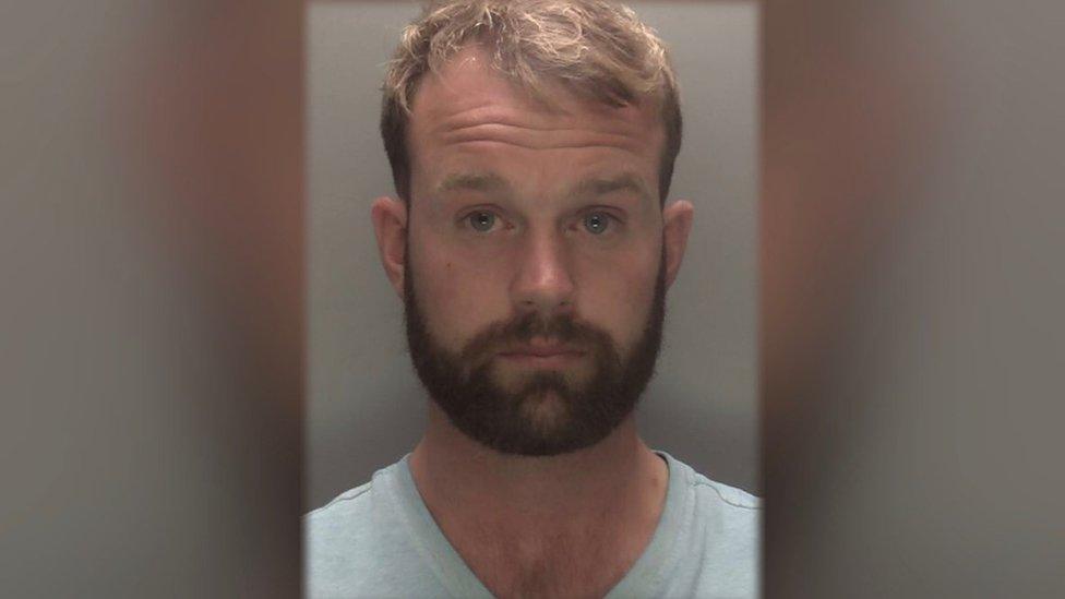 Man with brown hair and beard wearing light-coloured shirt in police mugshot