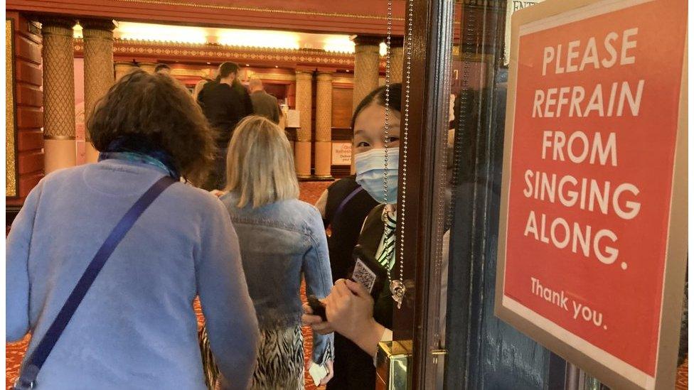 Theatre-goers walk past a sign in the theatre entrance that reads "Please refrain from singing along".