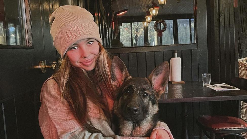 Liv holding her dog, wearing a pink "DKNY" hat and sweater. The background shows green wooden panels with a mirror reflecting the lights on the ceiling.