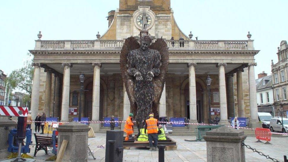 Human figure made up of knives in front of stone-pillared All Saints Church piazza