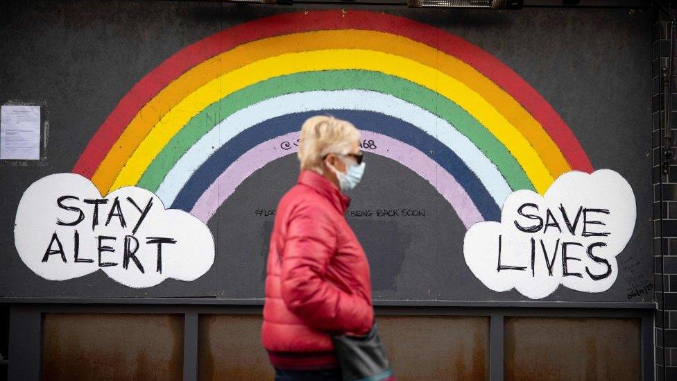 A woman wearing a face mask, sunglasses and a red coat walks past a rainbow mural on a wall which reads "Stay alert save lives".