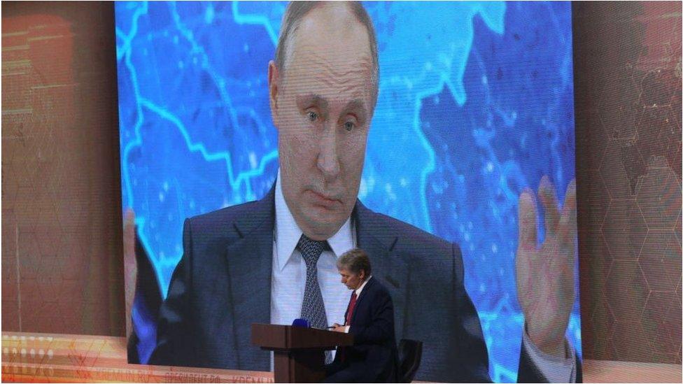 Dmitry Peskov in front of a screen showing President Putin