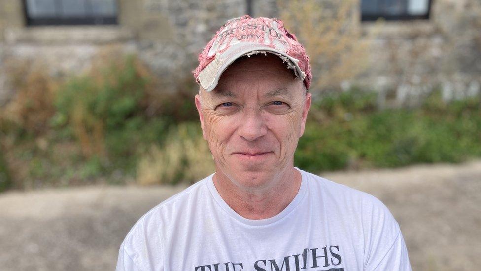 Gary Goodwin smiles at the camera, wearing a cap and a white T-shirt