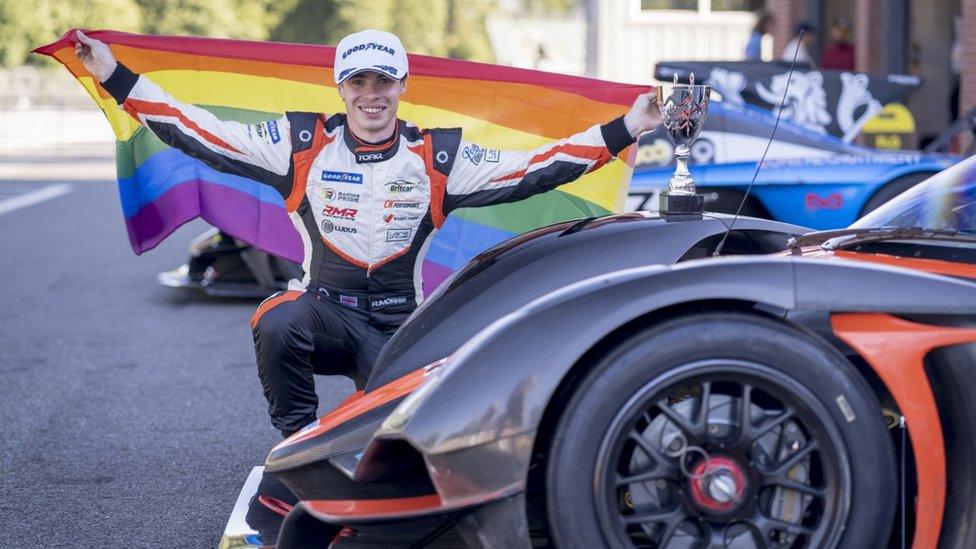 Richard Morris holding an LGBT flag by a trophy and racing car