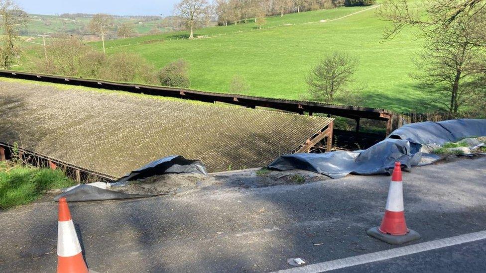 landslip on the side of the road with cones lining the road. There is a large green field in the background