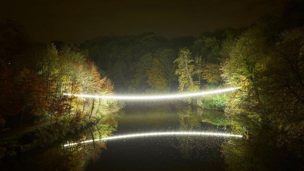 A string of bright, white light hangs over a still river, held on either side in a forest of autumnal trees.