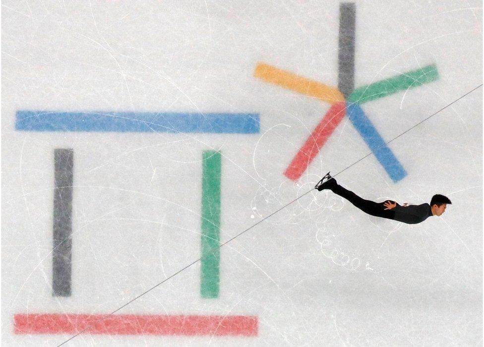 Skater Nathan Chen is seen with arms outstretched with coloured Olympic signs in the ice beneath him