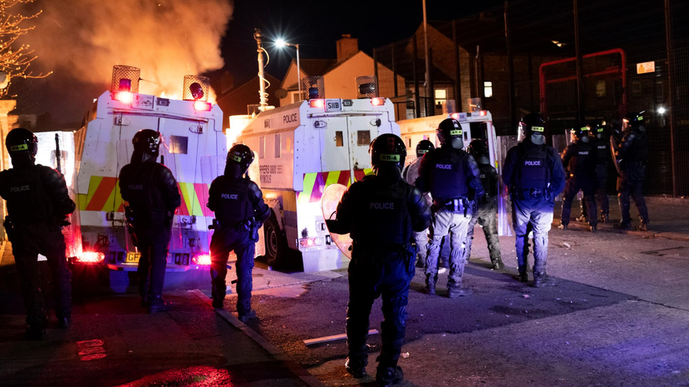 Police in riot gear watch as a car burns in front of police Land Rovers in Belfast