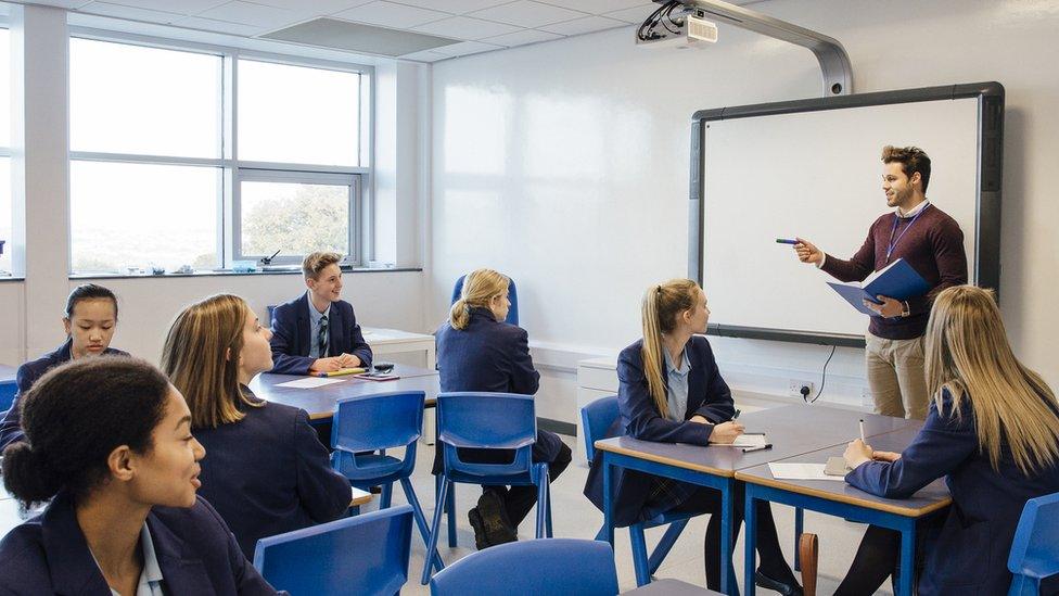 A teacher stands in front of a white board in a classroom