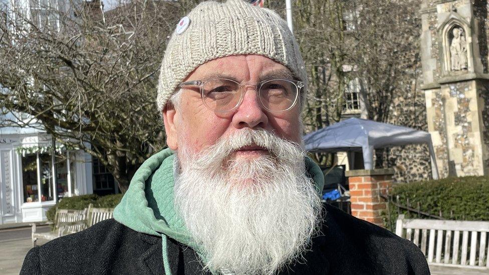 Man with long white beard and glasses stands in a street