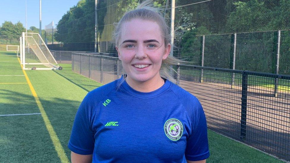 Kerry Brown on the Sion Swifts' home pitch, wearing their blue training top. She's stood by a yellow touchline, which disappears into the distance, with a set of goal posts visible side-on in the background. We can also see the wire fence surrounding the pitch and what looks like parkland - bushes and trees - just beyond it.