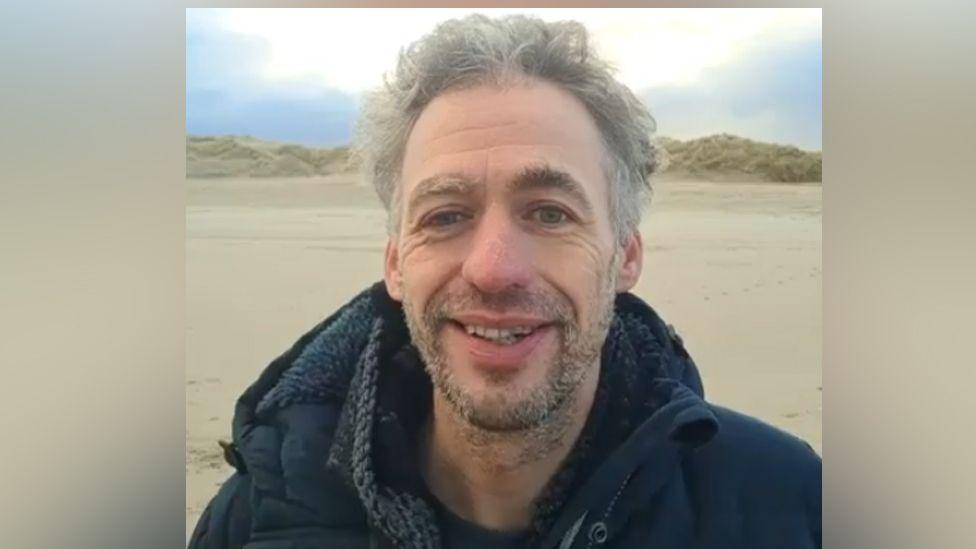 A head and shoulders image of Gert-Jan Van Der Kooij, wearing a navy hooded top standing on a sandy beach. There are dunes in the background. He has a slight beard.