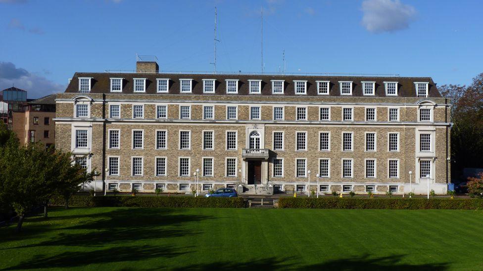 Shire Hall is a large, brick building with four stories and many windows. It is set in a large lawned area with hedges. There is a modern building behind it, to the left of the image.