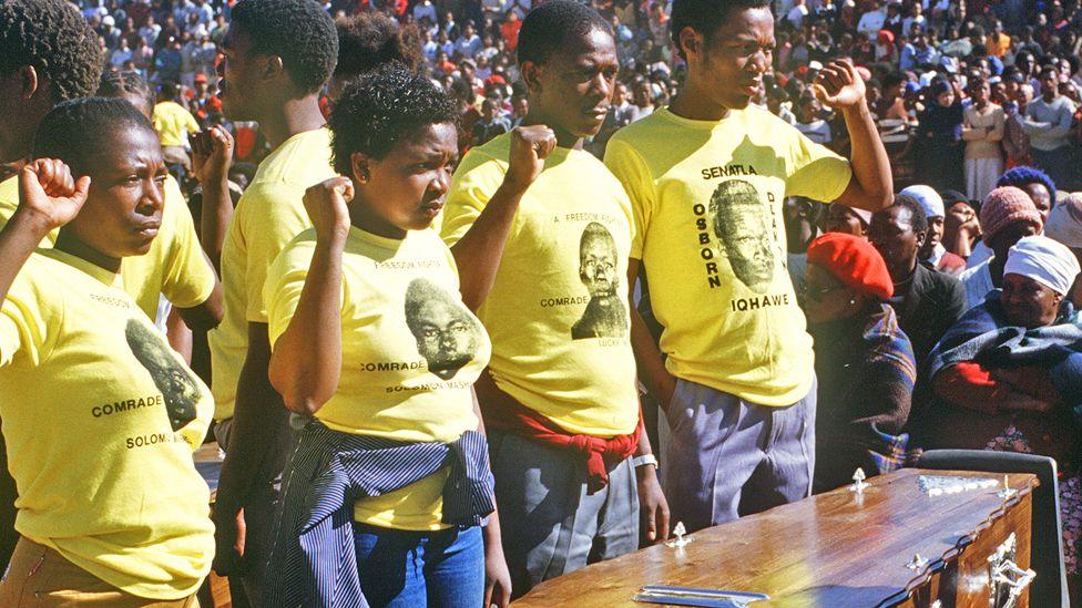 Young ANC activists in yellow T-shirts with their fists in the air next to a coffin of a fellow activist on 10 July 1985 in Duduza township, near Johannesburg