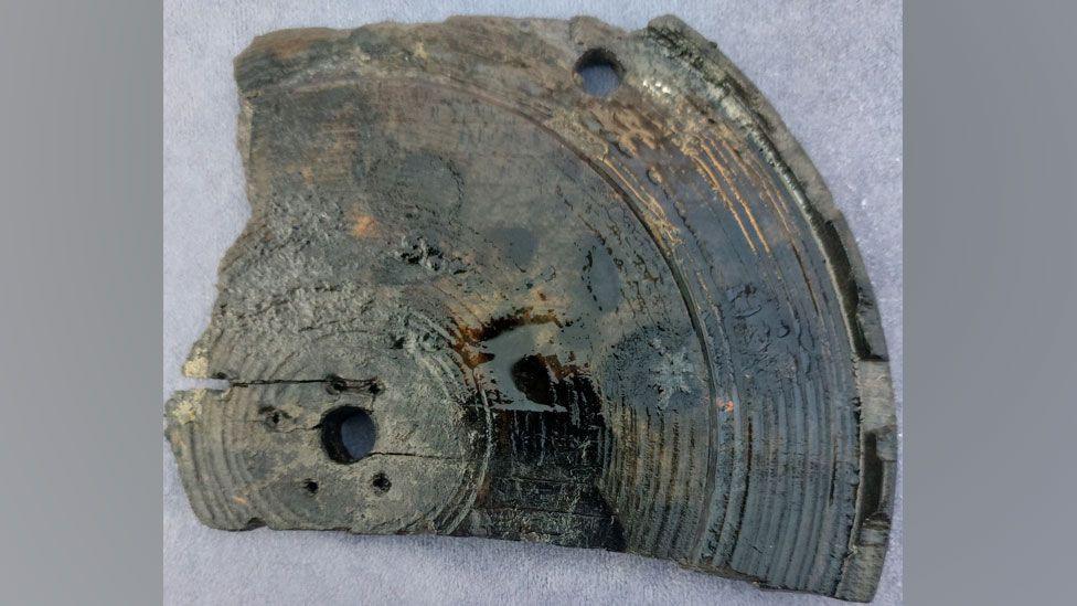 About a quarter of a round 17th Century wooden lantern base. In the middle is a large hole, surrounded by five smaller holes. The hole is surrounded by concentric circles until it reaches its raised edge.