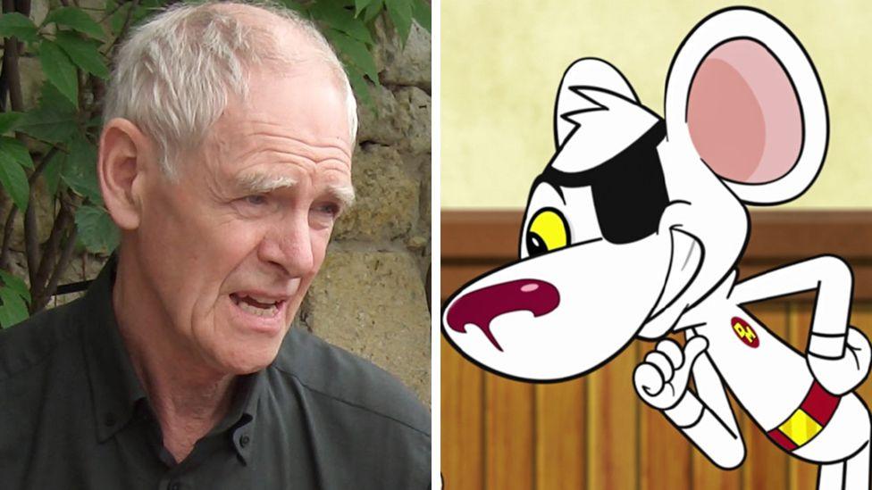 Brian Trueman, with white thinning hair and a black shirt, sits in fron tof a wall with greenery growing up it, alongside a cartoon of Danger Mouse, a white mouse with a red nose and big ears wearing a black eye patch and a red and yellow badge and belt