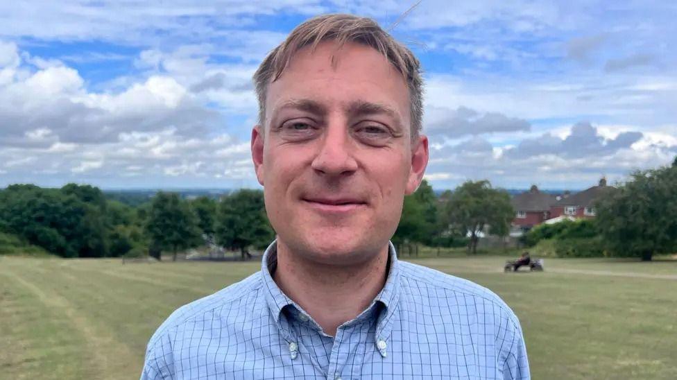 Olly Glover MP smiles for the camera, wearing a checked blue shirt. A neatly cut field is in the background behind him. 