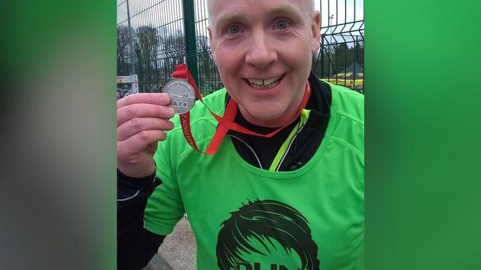 Larry at the finish line of a half marathon. He is wearing a green t shirt and is holding his medal up to the camera