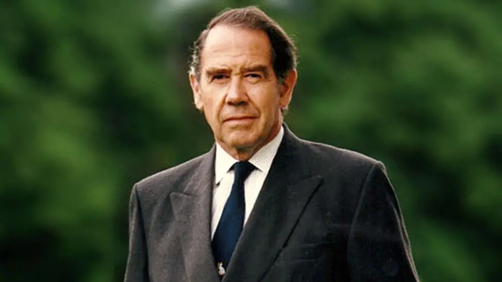 Sir Terence walks towards the camera looking at the lens, with one eyebrow slightly raised. We can just see him from the chest upwards. He wears a grey suit jacket, white shirt and blue tie. He has short dark hair and is clean shaven.