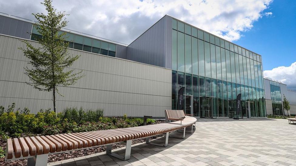 Large leisure centre building it is in a series of connecting blocks. The main entrance has a glass front with paving and shrubs.