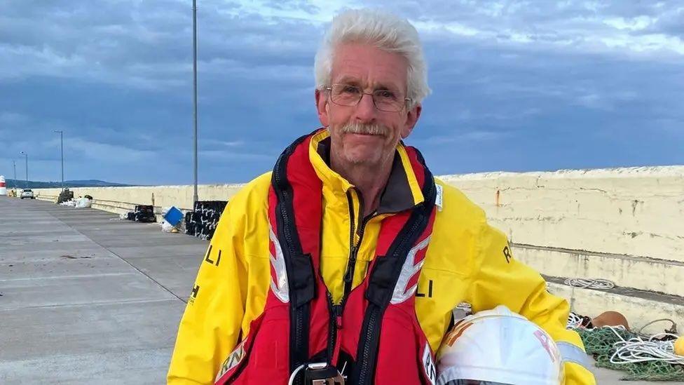 Mike Keggen a man with white hair, and a moustache wears glasses and a bright yellow RNLI coat and lifejacket and holds his helmet on the harbourside.