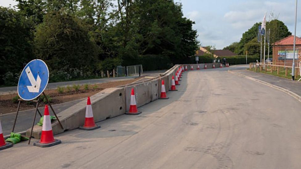 Concrete crash barriers along the side of a road with orange and white traffic cones in front of it and a blue road sign with arrow